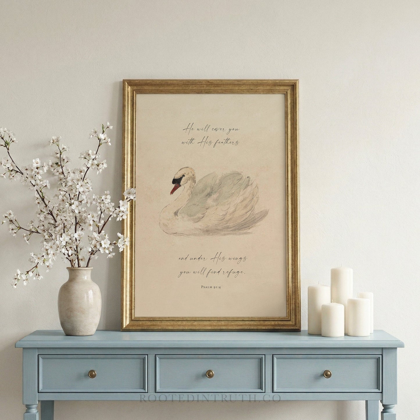 a framed print of a swan on a blue table, accompanied by a vase of white flowers and three white candles.