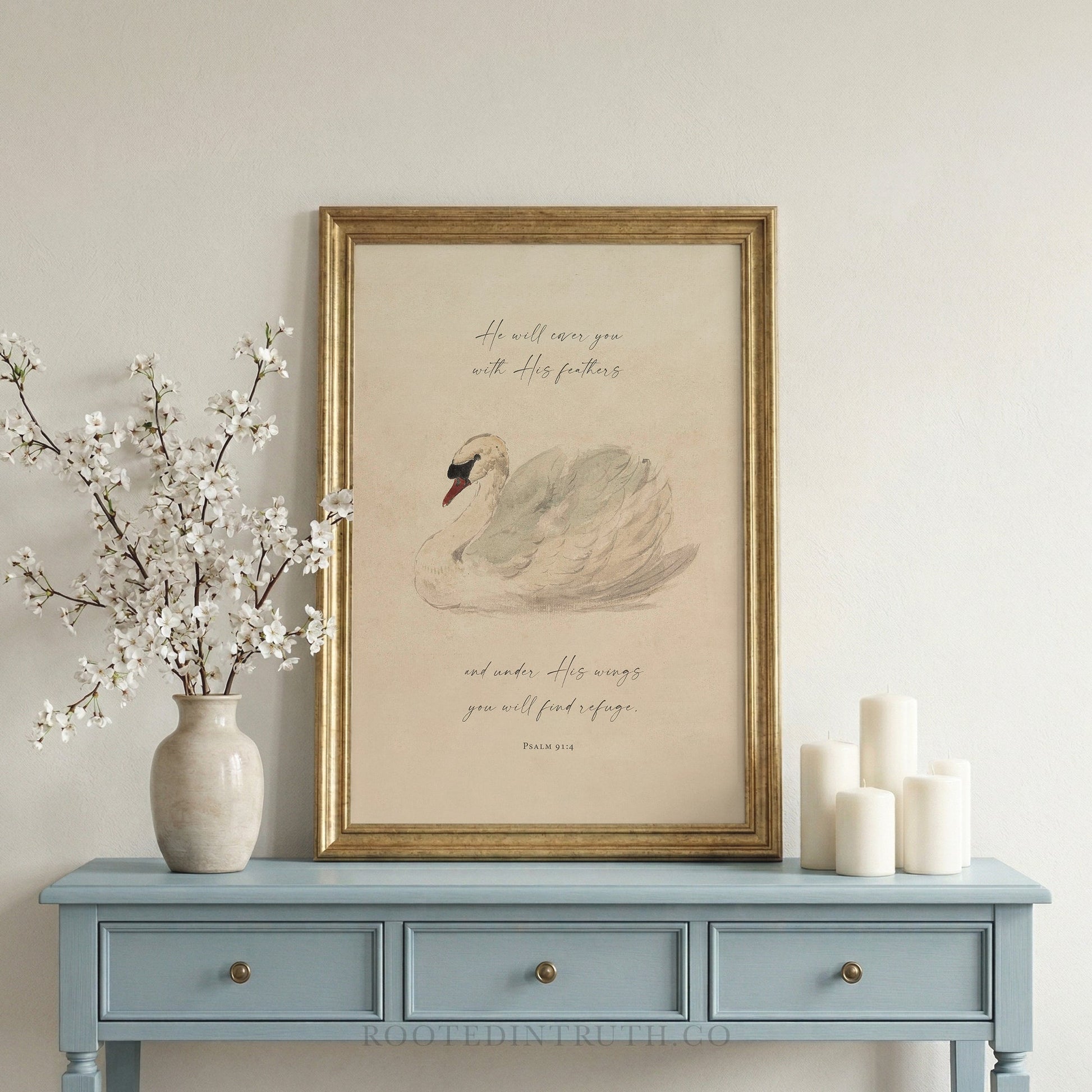a framed print of a swan on a blue table, accompanied by a vase of white flowers and three white candles.