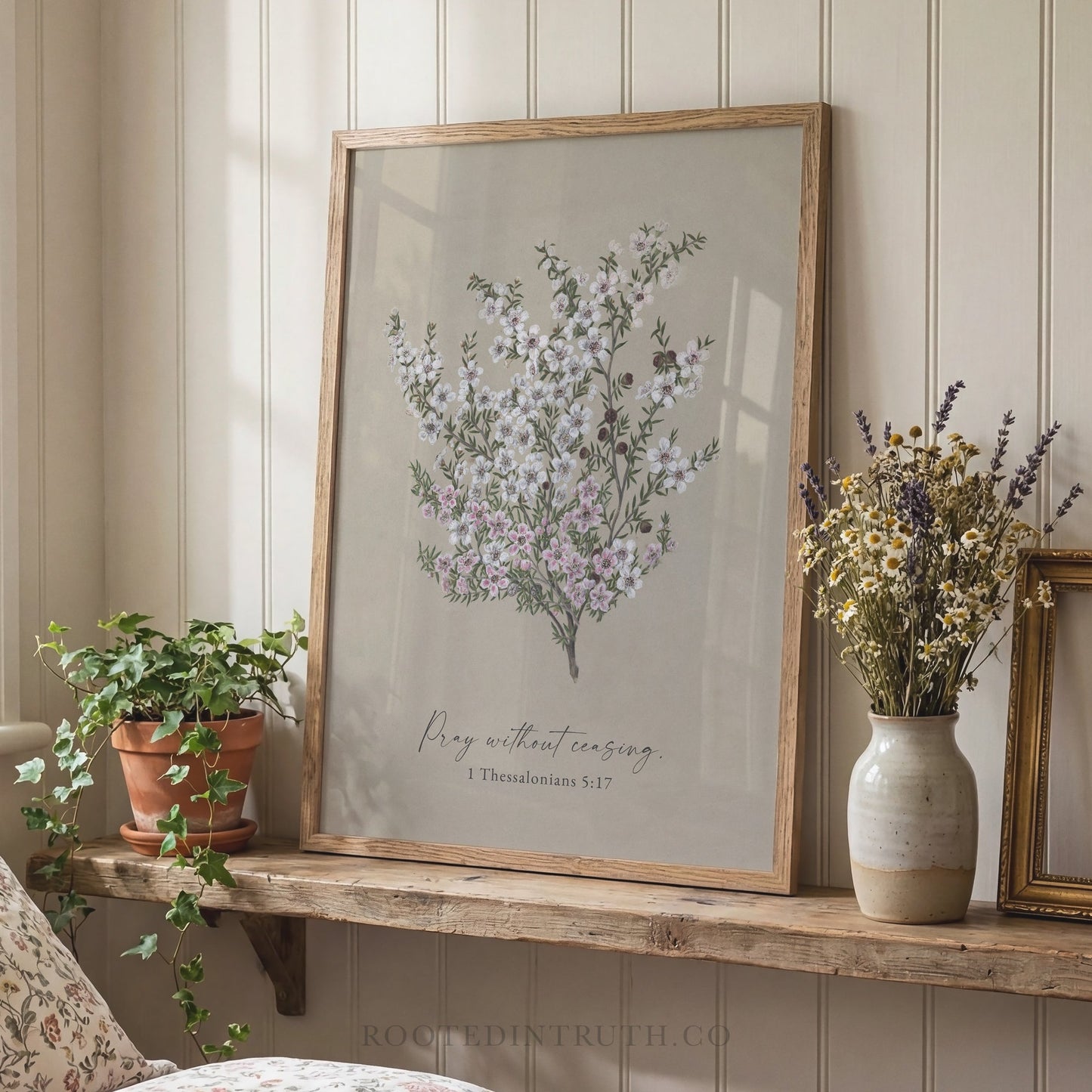 a framed print of a floral arrangement with the text "Pray without ceasing" and "1 Thessalonians 5:17" below it, placed on a wooden shelf next to a vase of flowers and a potted plant.