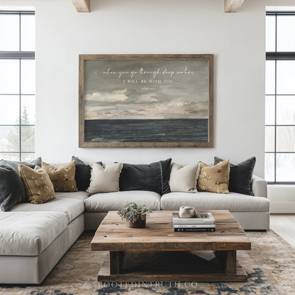 a living room with a large, framed painting of a seascape hanging on the wall above a gray sectional sofa. The sofa is adorned with throw pillows in various shades of brown and beige. In front of the sofa, there is a wooden coffee table with a plant on top. The room has large windows that allow natural light to fill the space, and the overall decor has a modern and minimalist aesthetic.