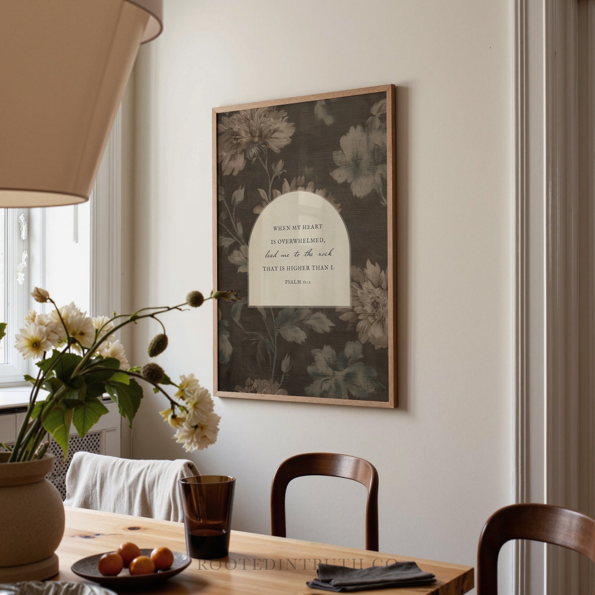 a framed print hanging on a wall above a wooden dining table with a vase of flowers and a plate of fruit.