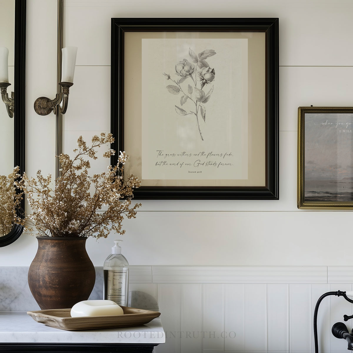 a bathroom setting with a framed print of a flower on the wall, a vase of dried flowers, a soap dispenser, and a mirror.