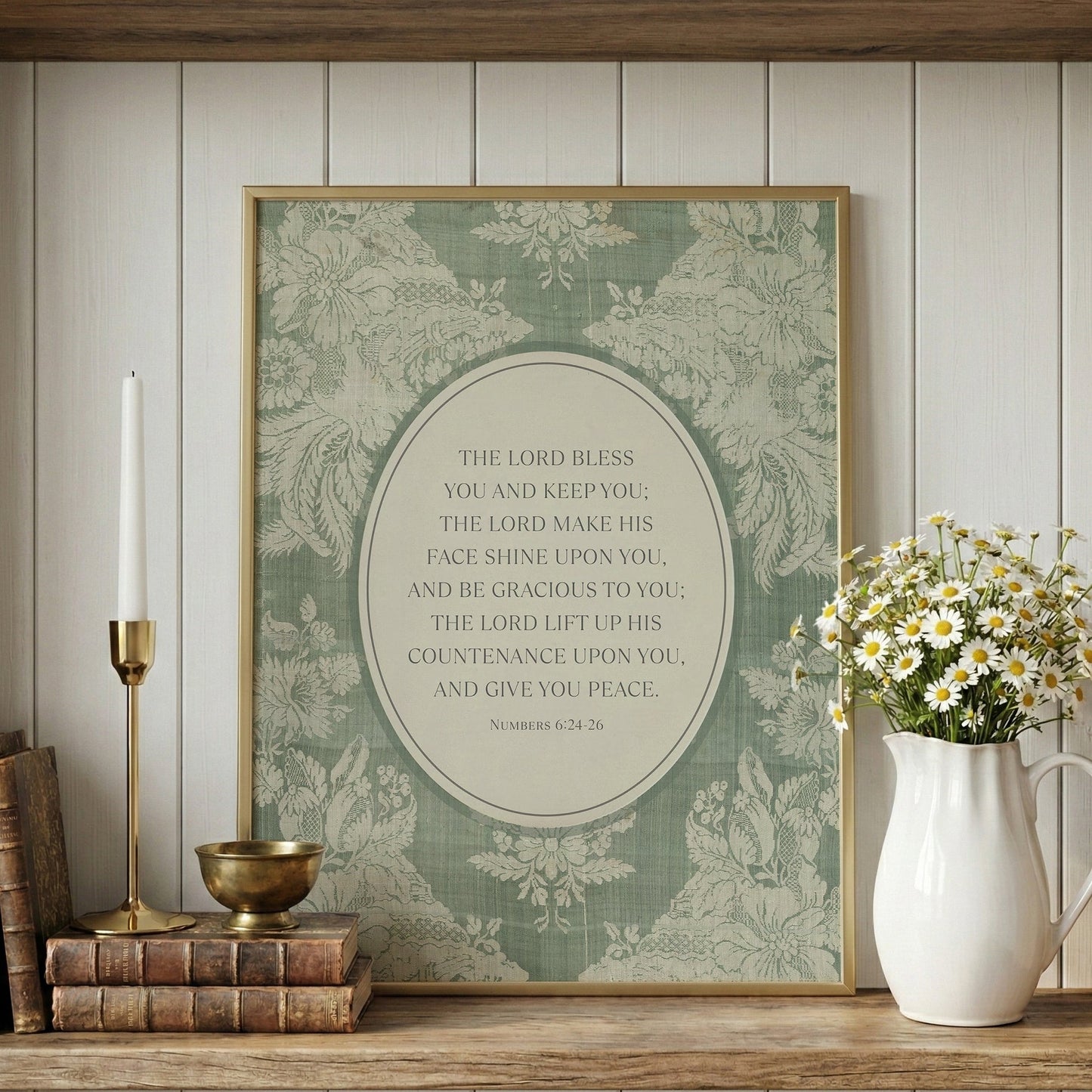 a framed print with a religious text, a white vase with white flowers, a stack of books, and a brass candlestick on a wooden shelf.