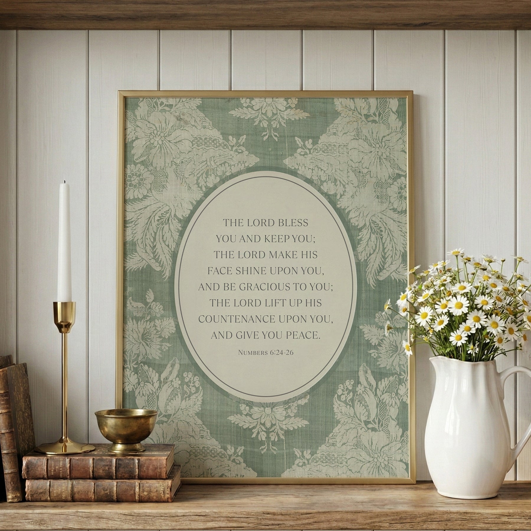a framed print with a religious text, a white vase with white flowers, a stack of books, and a brass candlestick on a wooden shelf.