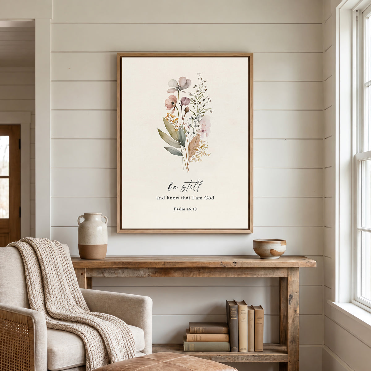 a cozy living room with a wooden console table, a beige armchair, and a framed print of a floral design above the table. The print includes a Bible verse and a quote from Psalm 46:10.