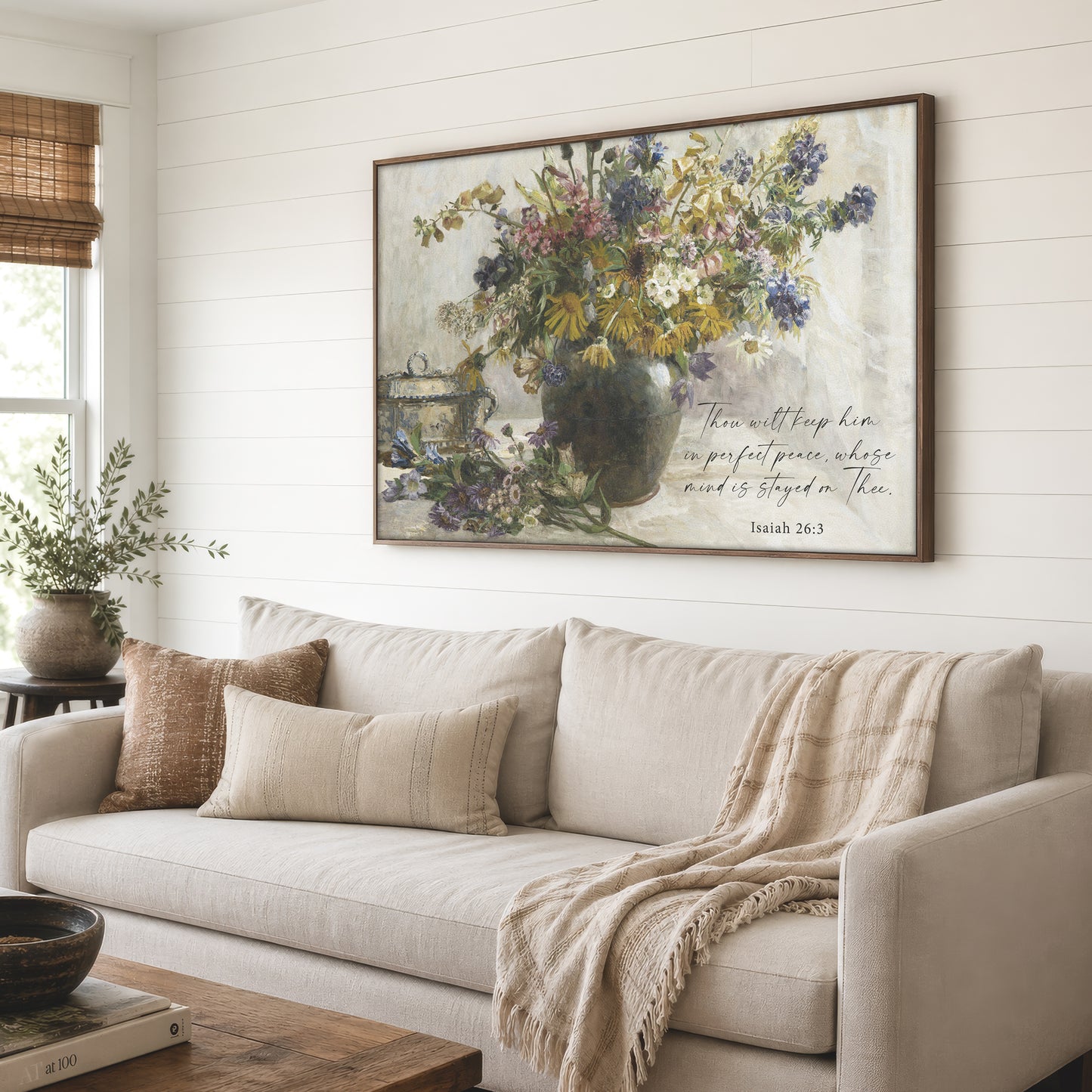 a large framed painting of a vase filled with flowers, hanging on a wall above a beige sofa with a throw blanket draped over it.