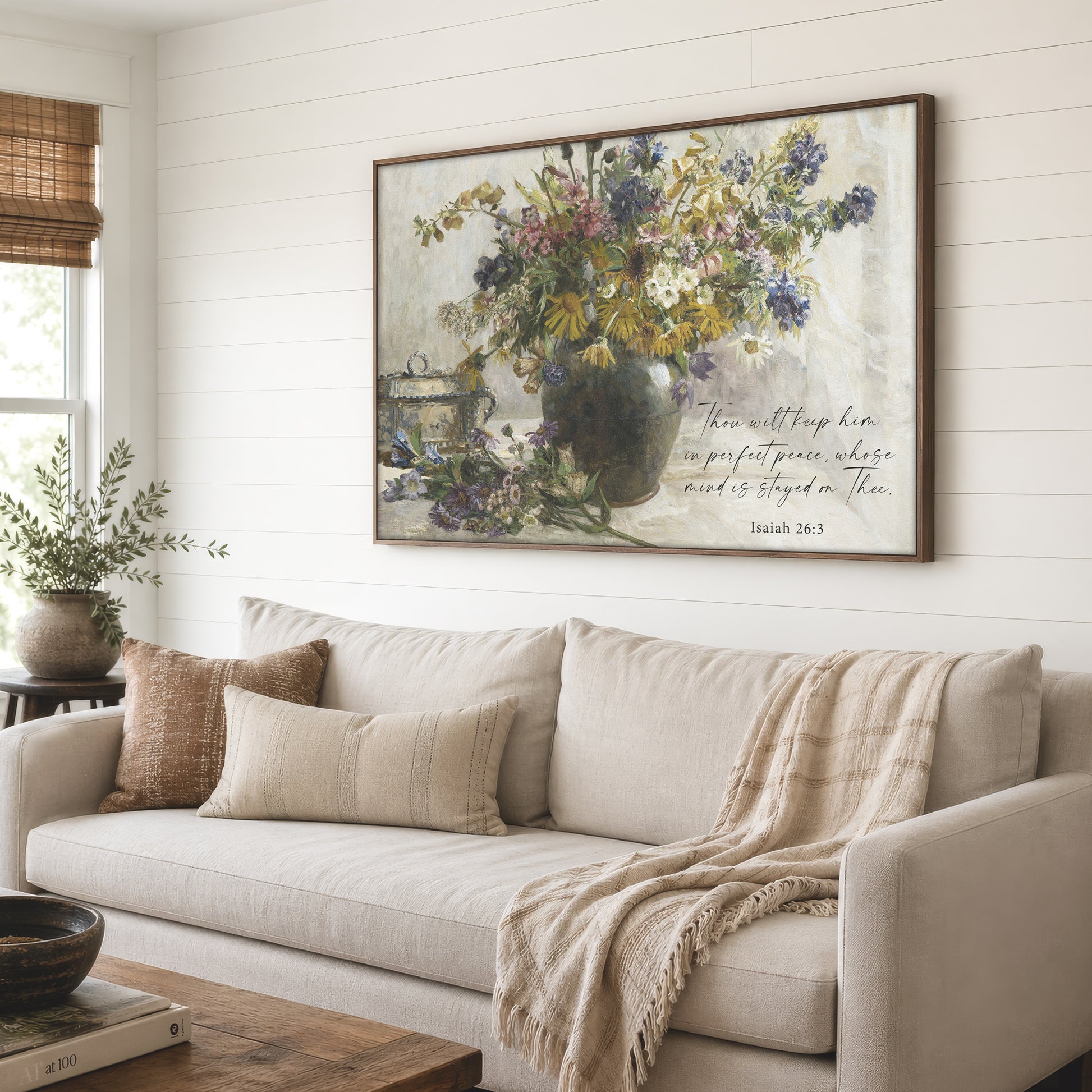 a large framed painting of a vase filled with flowers, hanging on a wall above a beige sofa with a throw blanket draped over it.
