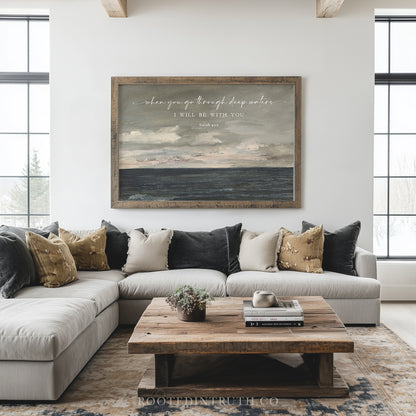 a living room with a large, framed painting of a seascape hanging on the wall above a gray sectional sofa. The sofa is adorned with throw pillows in various shades of brown and beige. In front of the sofa, there is a wooden coffee table with a plant on top. The room has large windows that allow natural light to fill the space, and the overall decor has a modern and minimalist aesthetic.