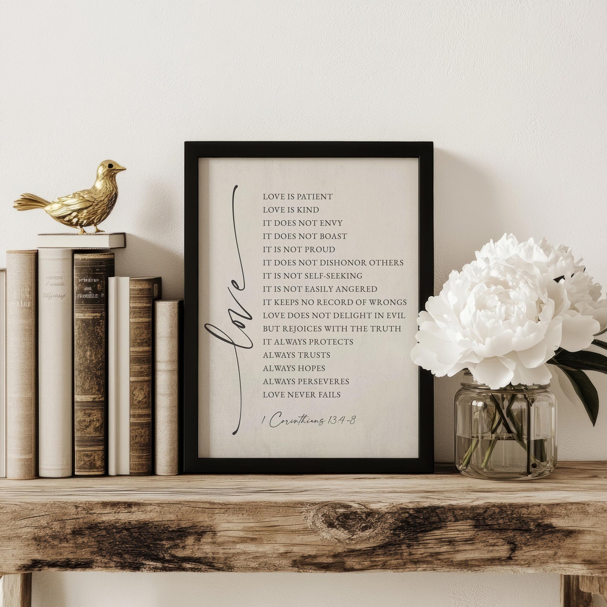 A framed print displaying a list of values and beliefs, including "Love is patient, love is kind, love does not boast, love does not envy, love is not disrespectful, love is not self-seeking, love is not keeping no record of wrongs, love always protects, love always hopes, and love never fails." The print is placed on a wooden shelf next to a vase of white flowers, with a gold bird figurine nearby.