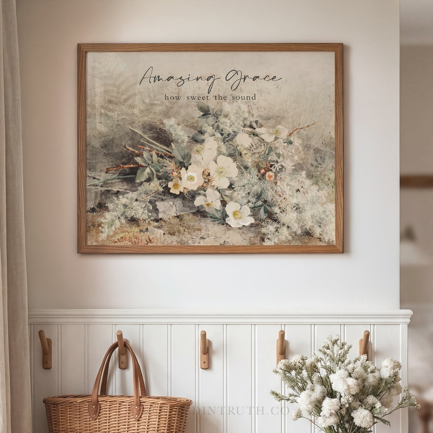a framed print of a floral arrangement hanging on a wall, with a wicker basket and a vase of white flowers placed below it.