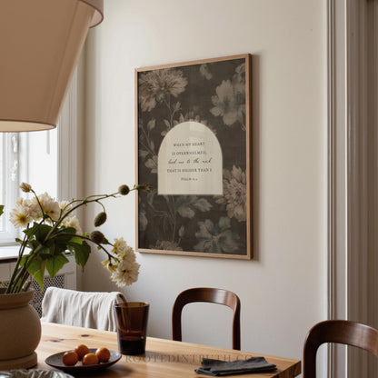 a framed print hanging on a wall above a wooden dining table with a vase of flowers and a plate of fruit.