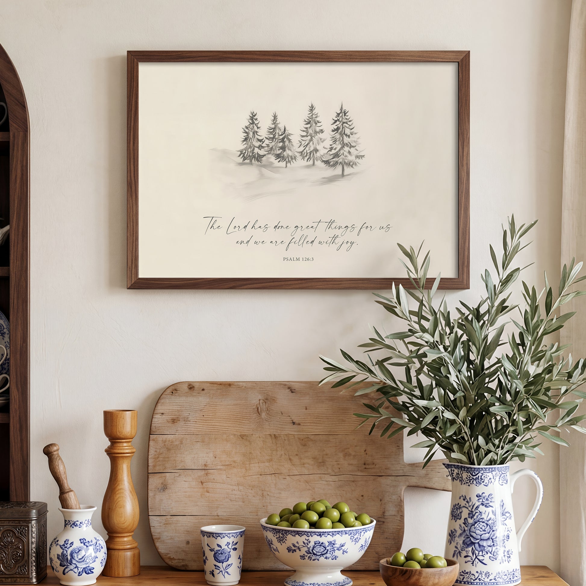 a wooden shelf with various decorative items, including a framed print of a winter landscape, a vase of green leaves, a bowl of fruit, and a wooden tray.