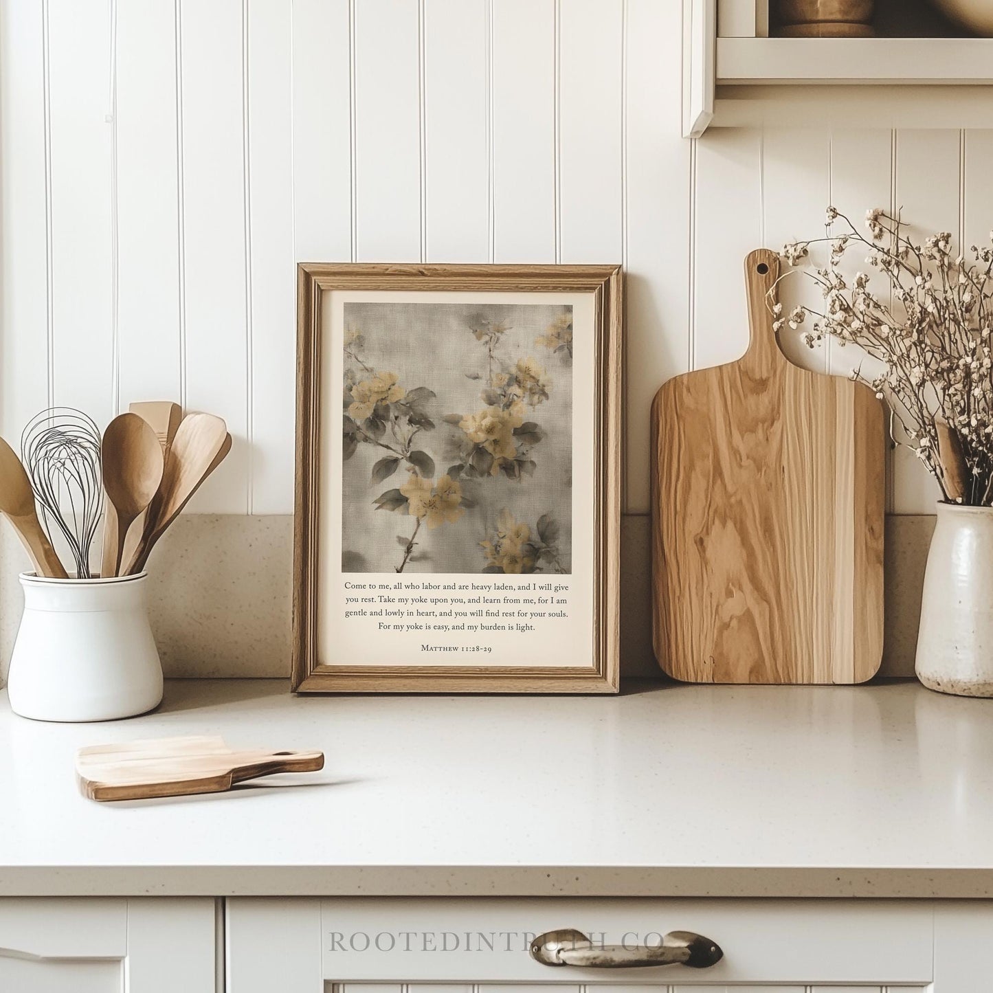 a framed print of a floral print on a white wall, accompanied by a wooden cutting board, a white vase with flowers, and a wooden spoon holder.