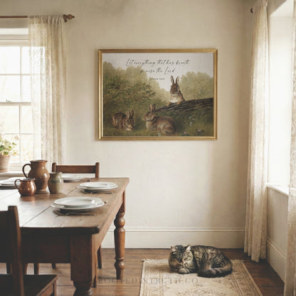 A cozy dining room with a wooden table set for two, featuring plates, silverware, and a vase. Above the table is a framed painting of three rabbits in a lush green forest. A cat is curled up on the floor in front of the table.